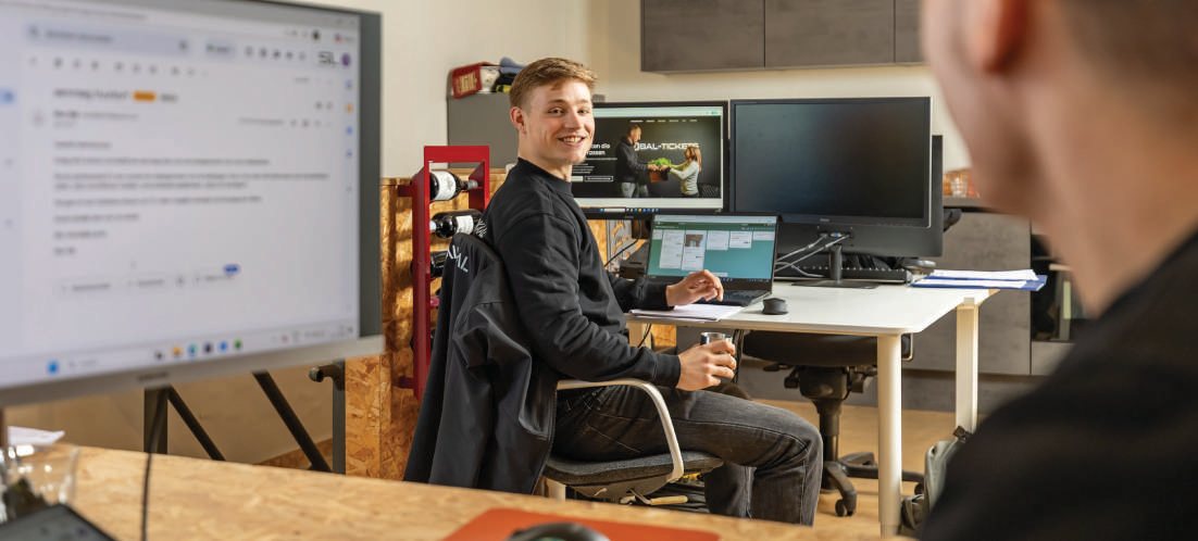 Teamlid aan een bureau met meerdere beeldschermen, omkijkend met een glimlach